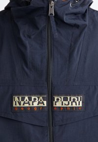Marineblå zip-up jakke med hætte, fremtrædende "NAPA PIJRI" logo badge i creme og orange, med en tekstureret stoffinish.
