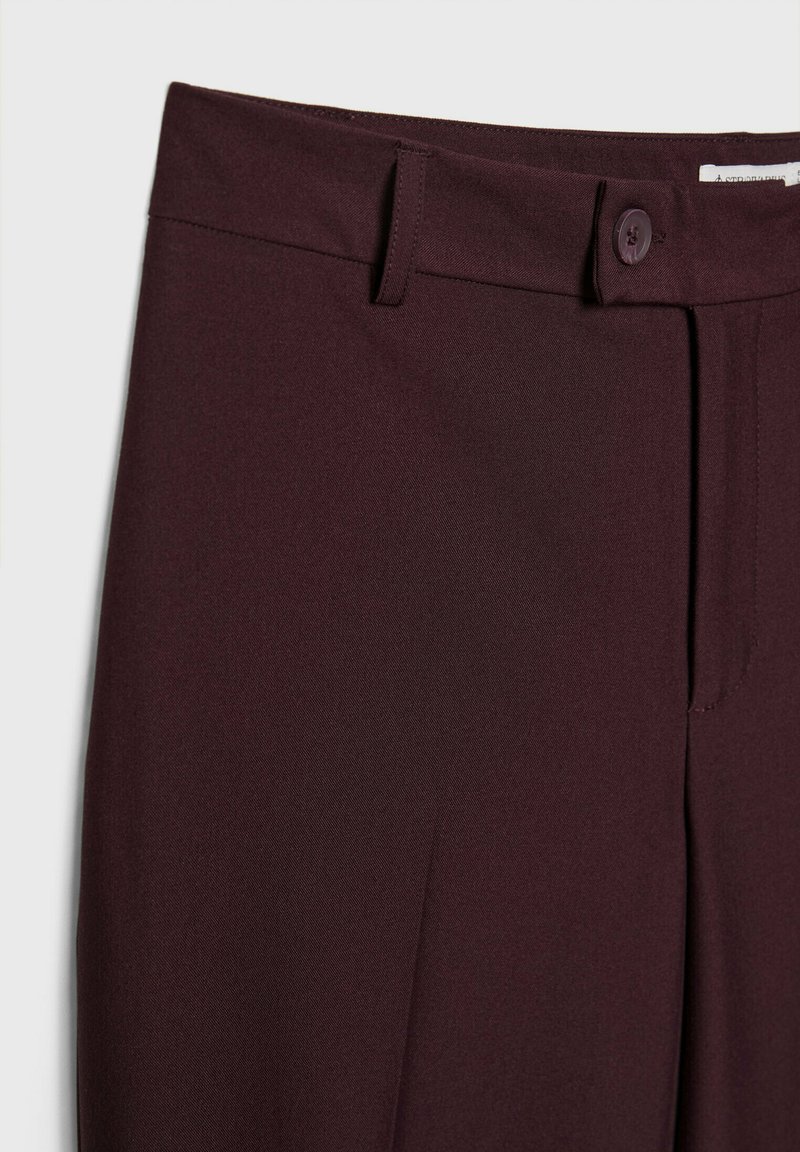 Pantaloni bordeaux realizzati in un tessuto liscio e testurizzato, con chiusura a un bottone e passanti per la cintura, progettati con una vestibilità su misura e dettaglio di piega.