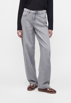 Person iført løse lysegrå jeans og mørkerøde sneakers med beige detaljer, stående foran en ensfarvet hvid baggrund.