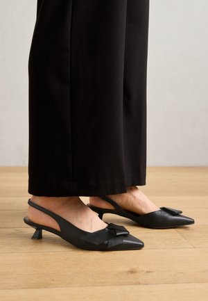 Scarpe slingback in pelle nera con punta affilata, caratterizzate da un dettaglio a fiocco sulla tomaia e un tacco basso a blocco. Posate su un pavimento in legno.