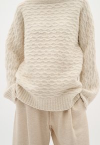 Strickpullover in hellbeige mit strukturiertem wellenförmigem Muster; gerippter Halsausschnitt und Saum. Kombiniert mit hellbeigen Jogginghosen mit glatter Oberfläche.