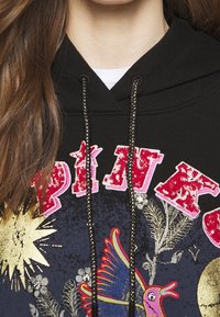 Svart huvtröja med rosa och gyllene grafiska tryck, texturerade dekorationer och metalliska snoddar. Designen inkluderar fåglar och himmelska motiv.