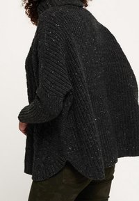 Krullend haar persoon in een losse, donkergrijze, ribbelgebreide trui met hoge kraag en donkere broek, gezien vanaf de achterkant.