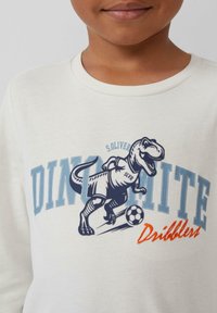 Witte lange mouwen T-shirt met een grafiek van een dinosauruss die voetbalt, met blauwe en oranje tekstaccenten. Zachte katoen stof.