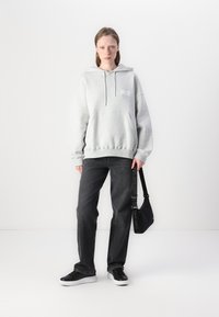 Grå hettegenser med frontlomme, kombinert med løse svarte jeans og svarte sneakers. Modellen holder en svart veske med logo detaljer.