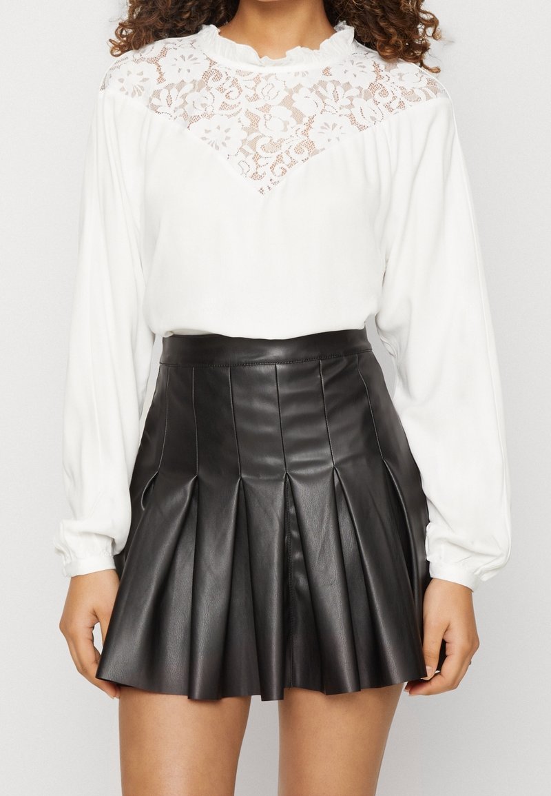 Blouse blanche à manches longues avec panneau en dentelle et col volanté, associée à une jupe plissée en cuir noir taille haute.