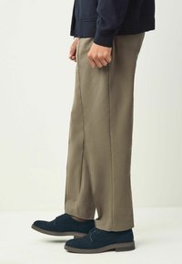 Pantalons habillés beige à coupe droite, associés à des chaussures en suede bleu marine avec lacets et semelles marron, sur un arrière-plan neutre.