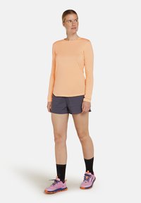 Kvinna som bär en persikofärgad långärmad skjorta, mörkgrå shorts, svarta strumpor och rosa löparskor, stående mot vit bakgrund.