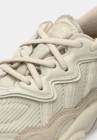 Närbild av beige Adidas-sneaker med texturerad tyg, läderöverdrag och snören som korsar den vadderade tungan med Adidas-logotyp.