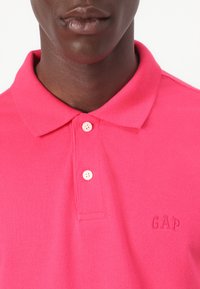 Lyserød polo shirt i en tekstureret stof, med krave og to knapstolper. Broderet "GAP" logo i matchende farve.