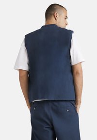 Chaleco sin mangas de color azul marino hecho de una tela suave, con un escote redondeado y un corte recto. Combinado con una camisa blanca y pantalones cortos azul marino.