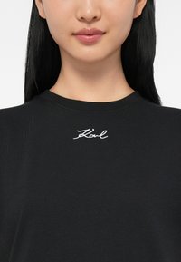 Κοντινό πλάνο ενός ατόμου που φοράει ένα μαύρο πουκάμισο με το "Karl" κεντημένο σε άσπρη γραφή κάτω από τον λαιμό.