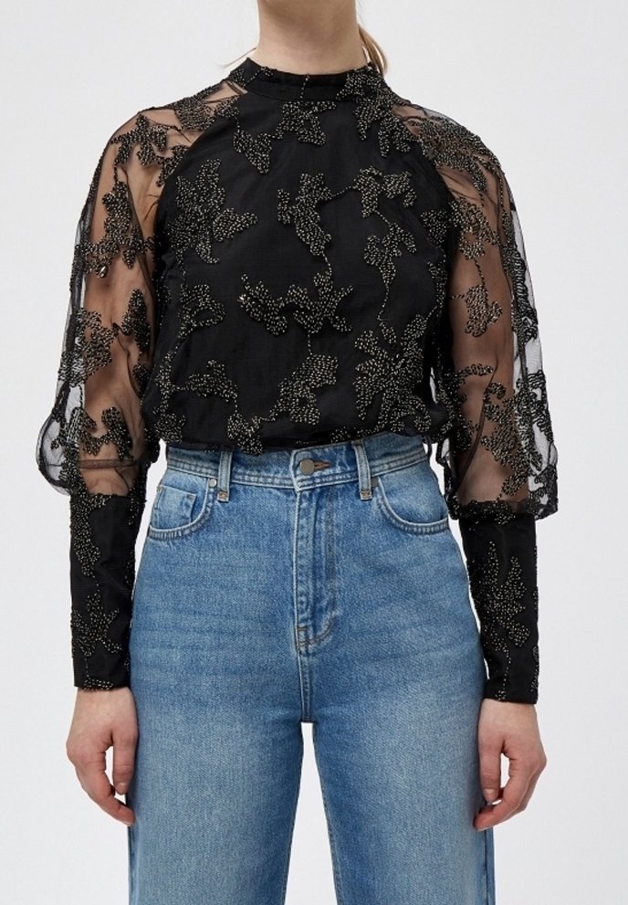 Blouse noire transparente avec broderie florale en perles portée avec un jean bleu taille haute, montrant des manches longues détaillées et un col haut.