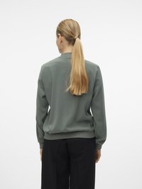 Femme aux longs cheveux blonds attachés en queue de cheval, portant une veste bomber verte et un pantalon noir, debout de dos sur un fond blanc.