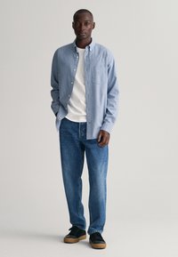 Camisa azul clara de botones sobre una camiseta blanca, combinada con jeans azul oscuro y zapatillas negras con suelas marrones. Diseño sencillo, ajuste casual.