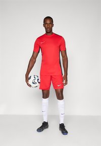Rød Nike sportsdrakt og shorts med svarte detaljer, kombinert med hvite sokker og svarte fotballsko, som holder en mønstret fotball.