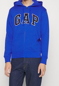 Sweat à capuche zippé bleu avec une poche avant, une capuche à cordon et le logo "GAP" en noir sur la poitrine. Confectionné en tissu doux.
