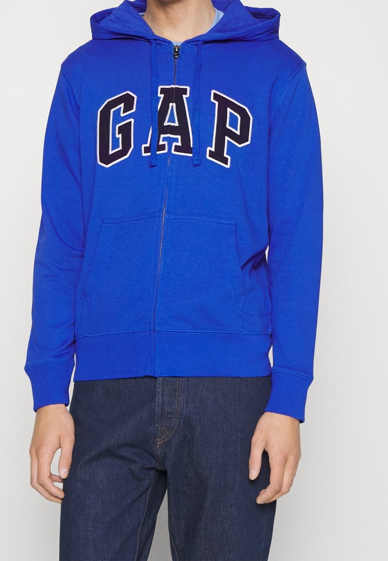Sweat à capuche zippé bleu avec une poche avant, une capuche à cordon et le logo "GAP" en noir sur la poitrine. Confectionné en tissu doux.