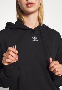 Černá bavlněná mikina s kapucí a šňůrkou, na přední straně s bílým vyšívaným logem Adidas na hrudi a žebrovanými manžetami. Hladký povrch.