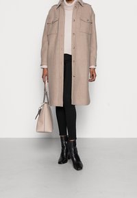 Persoon draagt een beige lange jas, lichtkleurige trui, zwarte broek, zwarte enkellaarsjes en houdt een beige handtas met kettingriem vast, tegen een witte achtergrond.