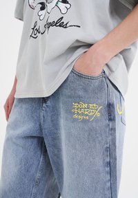 Camiseta gris de manga corta con estampado gráfico y texto, combinada con jeans de mezclilla azul claro que presentan un logotipo bordado en amarillo en el bolsillo.