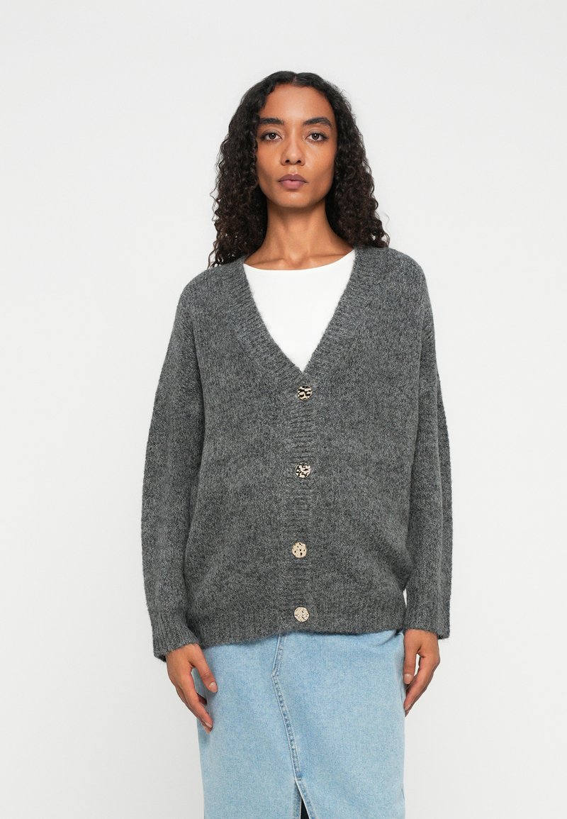 Kvinne med krøllete hår iført en grå knappet cardigan over en hvit topp og et lyseblått denimskjørt som står mot en ensfarget bakgrunn.