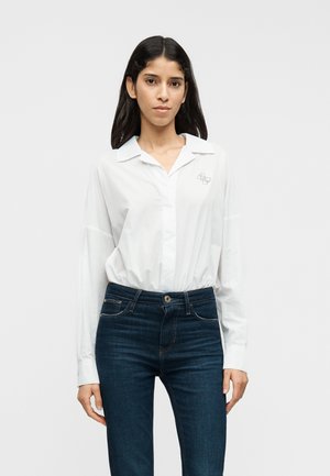 Hvid langærmet skjorte med knapper og krave, prydet med en lille broderet logo på venstre bryst og kombineret med mørkeblå jeans.