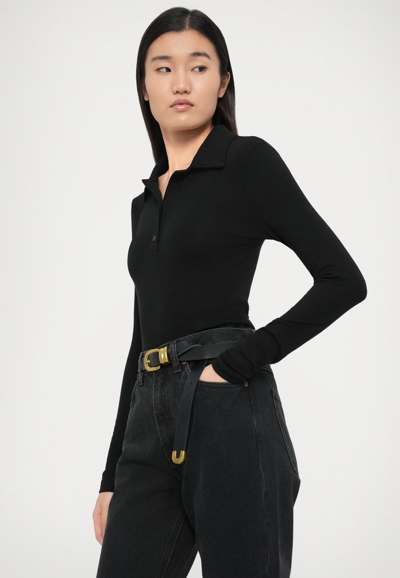 Jonge vrouw draagt een zwart lange mouwen polo, zwarte jeans met hoge taille en een zwarte riem met gouden gesp, poserend met één hand in de broekzak.