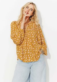 Femme blonde portant un blouse à fleurs moutarde à manches longues et un jean bleu clair pose avec un doigt près des lèvres contre un fond blanc.