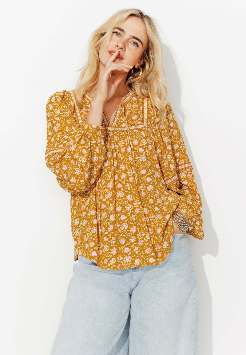 Femme blonde portant un blouse à fleurs moutarde à manches longues et un jean bleu clair pose avec un doigt près des lèvres contre un fond blanc.