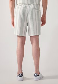 Shorts légers à rayures vertes et blanches. Taille élastique avec une petite poche arrière. Le mannequin porte des baskets blanches.