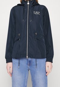 Mörkblå vindjacka med dragkedja framtill, justerbara dragskor, två sidofickor och en logotyp på bröstet. Har elastiska muddar.