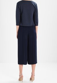 Haut bleu marine à pois avec manches trois-quarts, associé à un pantalon culotte taille haute. Tissu lisse ; chaussures à talons fines complètent la tenue.