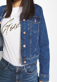 Modrá džínová bunda s měděnými knoflíky a dvěma náprsními kapsami. Nahoře přes bílou košili se zlatým vzorovaným motivem. Pod bundou jsou vidět přiléhavé džíny.