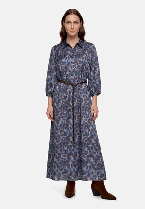 Femme portant une robe boutonnée à manches longues à motif bleu et gris, une fine ceinture marron et des bottines marron, debout face à l'avant.