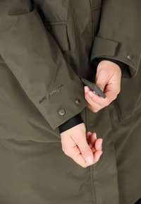 Olivgrön isolerad kappa med en mjuk textur, har tryckknappar på ärmarna och en vattentät etikett tryckt i svart.