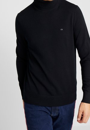 Homme portant un pull à col roulé noir avec un petit logo « ck » sur la poitrine, associé à un jean bleu sur un fond uni.