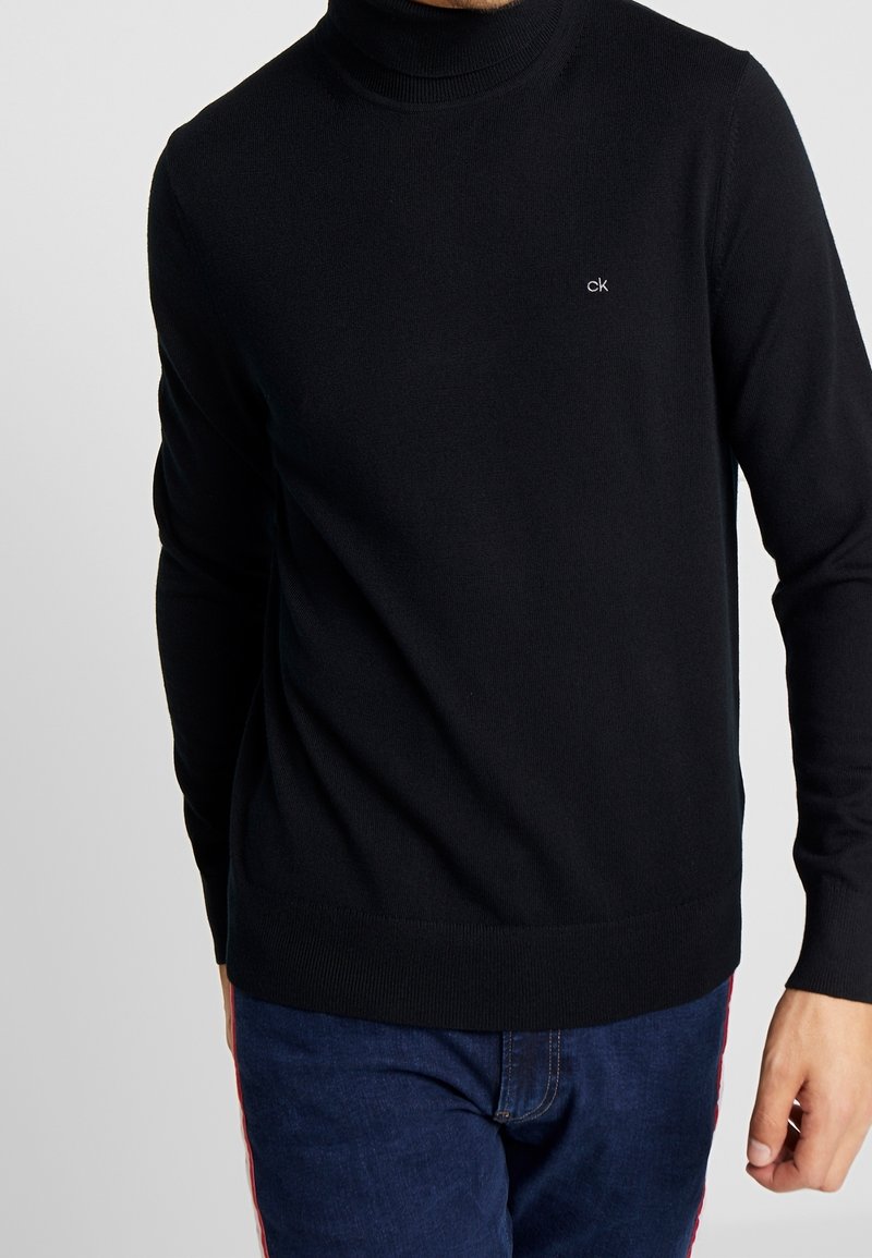 Homme portant un pull à col roulé noir avec un petit logo « ck » sur la poitrine, associé à un jean bleu sur un fond uni.