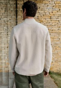 Light beige fleece jacket with a soft texture, featuring a traditional collar and subtle stitching details, paired with dark green pants.