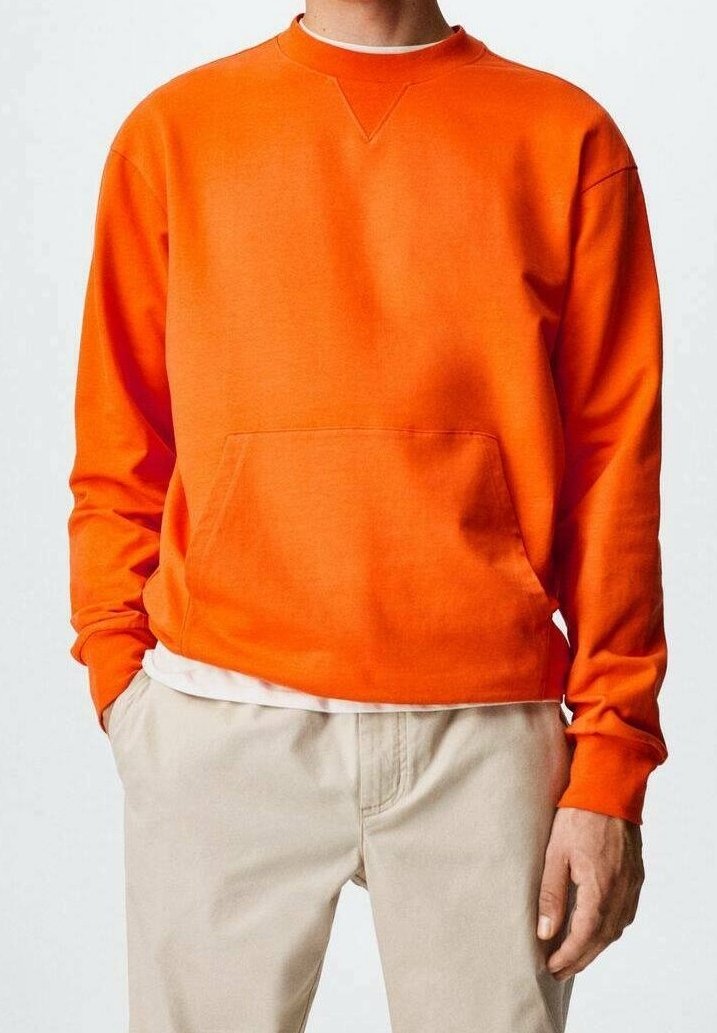 Personne portant un sweat-shirt à manches longues orange vif avec poche avant et pantalon beige, mains détendues sur les côtés, fond blanc.