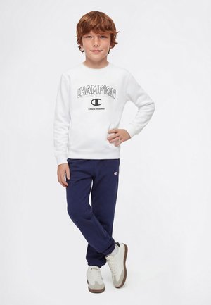 Niño con cabello rojo que lleva una sudadera blanca de Champion y pantalones deportivos azul marino, de pie con una mano en la cadera y las piernas cruzadas.