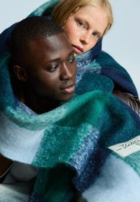 Wool-blend blanket featuring a textured finish, in shades of green, navy, and gray with varying patterns and a stitched label detail.