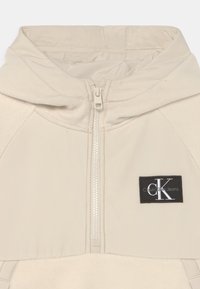Giacca con cappuccio beige con zip frontale, caratterizzata da una morbida texture in pile e da una patch nera con il logo Calvin Klein Jeans sul petto.