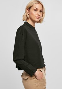 Kuttet svart langermet polo skjorte med knapper i halsen og ribbestrikket krage, kombinert med beige bukser. Glatt tekstur, avslappet passform.