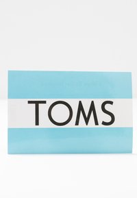 Boîte à chaussures bleu clair avec une bande blanche au centre affichant le nom de la marque en noir gras "TOMS" sur un fond blanc.