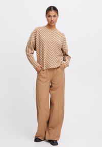 Pull en tricot à motif chevron marron avec des manches longues, associé à un pantalon large taille haute beige et des chaussures à semelle épaisse noires.