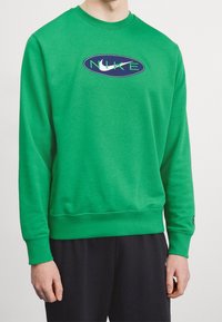 Zelená mikina se zaobleným výstřihem, žebrovanými manžetami a lemem. Na přední straně je modrý oválný logo s nápisem 'NIKE' v zelené barvě. Měkká textilie.