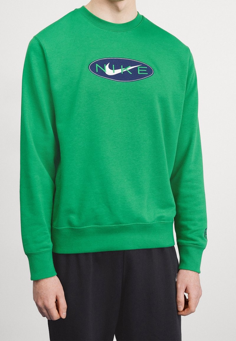 Zelená mikina se zaobleným výstřihem, žebrovanými manžetami a lemem. Na přední straně je modrý oválný logo s nápisem 'NIKE' v zelené barvě. Měkká textilie.