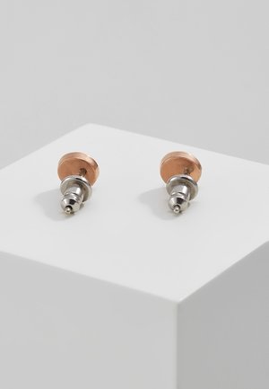 Een paar koperen en zilverkleurige metalen studs die op een witte kubus liggen tegen een lichtgrijze achtergrond.