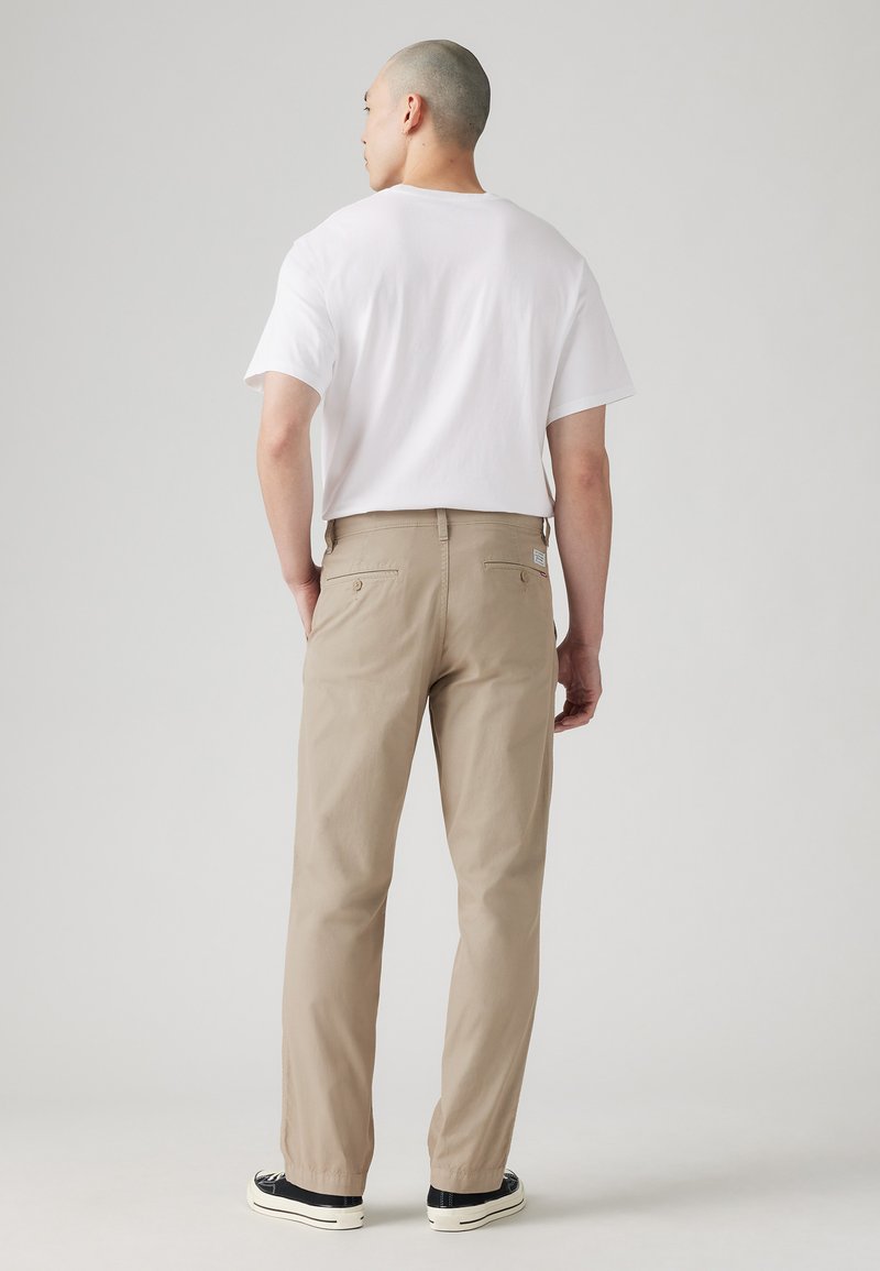 Pantalons beiges en tissu léger, dotés de deux poches arrière, portés avec un T-shirt blanc et des baskets noires.
